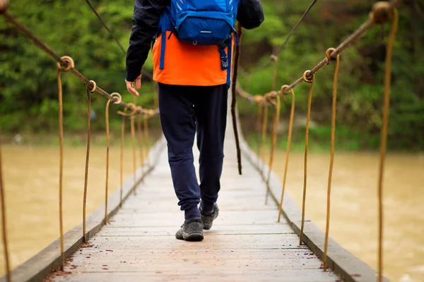 Hiking man crossing river in walking in balance on hinged bridge in nature landscape. Closeup of male hiker trekking shoes outdoors in forest balancing on bridge.