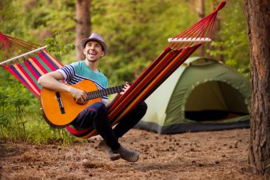 Gerçek bir rahatlama. Şapka lı yakışıklı genç adam hamakta yatarken gitar çalıyor , arka planda kamp