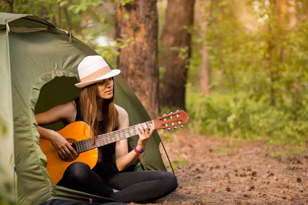 Chica turística en sombrero se sienta en la tienda y el concepto de tocar la guitarra. El turismo descansa en la naturaleza. Camping estilo de vida — Foto de Stock