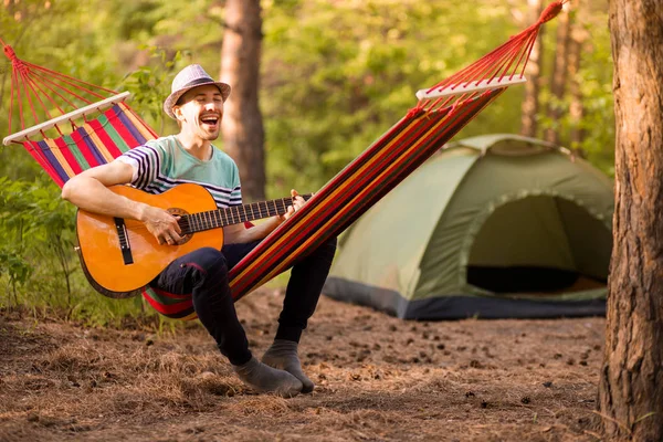 Joven caucásico hombre en sombrero jugar guitarra y balanceo en hamaca de fin de semana por la mañana en el campamento de descanso — Foto de Stock