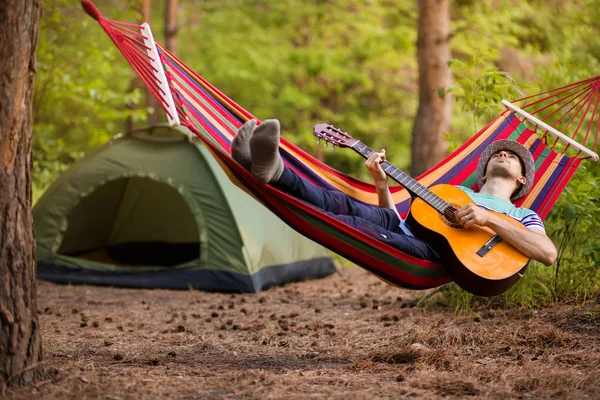 Real relaxation. Handsome young man in hat play guitar while lying in hammock , camping on background