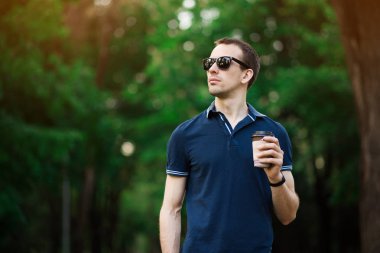 Güneş gözlüğü takan koyu saçlı çekici genç adam güneşli bir günde bir fincan kahve ile sokakta yürüyor. Rahat giysiler giyen sakallı hipster adam bir yaz hafta sonu rahatlatıcı sahiptir.