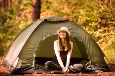 Şapkalı mutlu kadın gün batımı ışık gökyüzü altında orman etrafında çadırda boş ve özgürlük zevk oturur.