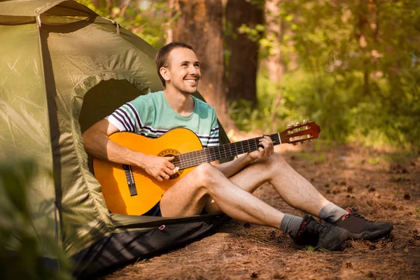 Happy young man camping and strum a guitar instrumental music to relax against background of forest sunset. Wilderness Travel lifestyle concept.