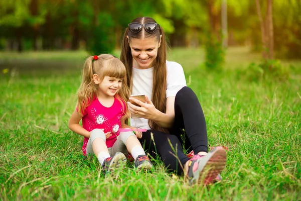 Χαρούμενα χαμογελαστά ευτυχισμένη μητέρα και η μικρή κόρη της κάθονται σε πράσινο γρασίδι στο καλοκαιρινό πάρκο και χρησιμοποιώντας ένα τηλέφωνο για να παρακολουθήσετε κινούμενα σχέδια, ένα κορίτσι δείχνει στην οθόνη το Σαββατοκύριακο — Φωτογραφία Αρχείου
