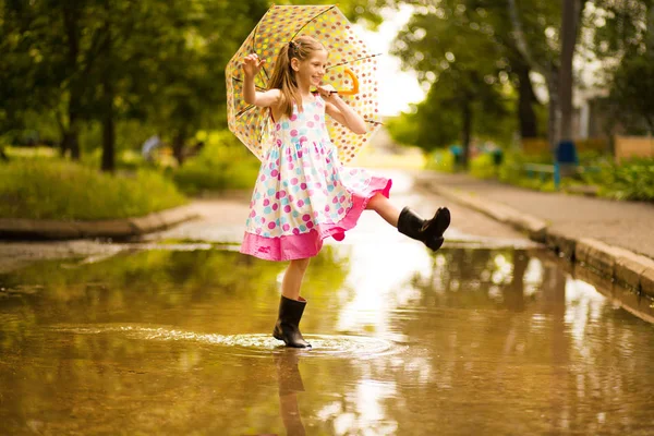Fröhliches lustiges Kindermädchen mit Regenschirm, das in Gummistiefeln und im Tupfen-Kleid auf Pfützen springt und lacht — Stockfoto