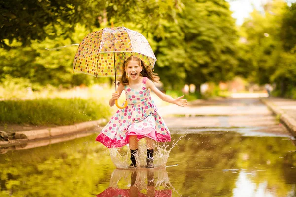 Menina garoto engraçado feliz com guarda-chuva pulando em poças em botas de borracha e em polka dot vestido e rindo — Fotografia de Stock