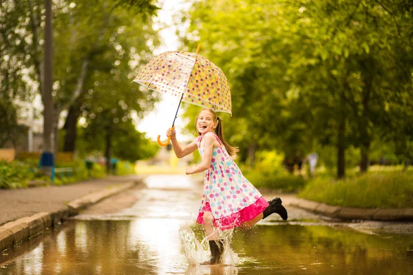 Menina garoto engraçado feliz com guarda-chuva pulando em poças em botas de borracha e em polka dot vestido e rindo — Fotografia de Stock