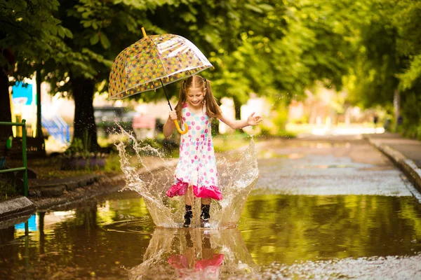 Feliz niña divertida con paraguas saltando sobre charcos en botas de goma y en vestido de lunares y riendo —  Fotos de Stock