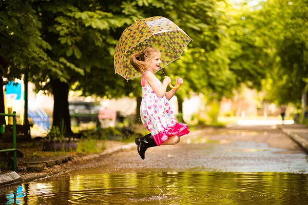 Menina garoto engraçado feliz com guarda-chuva pulando em poças em botas de borracha e em polka dot vestido e rindo — Fotografia de Stock