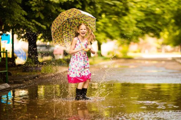 Menina garoto engraçado feliz com guarda-chuva pulando em poças em botas de borracha e em polka dot vestido e rindo — Fotografia de Stock