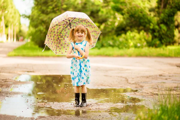 Happy child girl in dress with an umbrella and rubber boots in puddle on summer walk — Stock Photo, Image