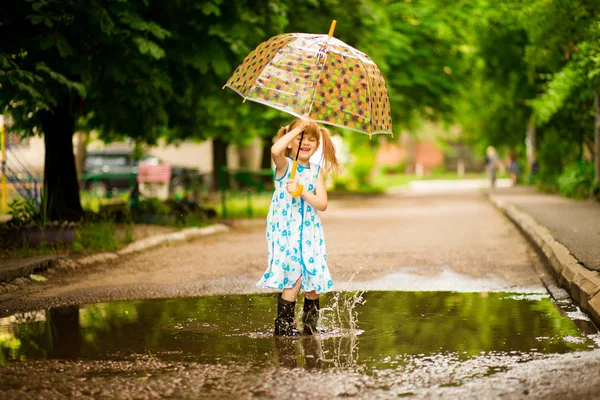 Feliz niña divertida con paraguas saltando sobre charcos en botas de goma y en vestido de lunares y riendo —  Fotos de Stock