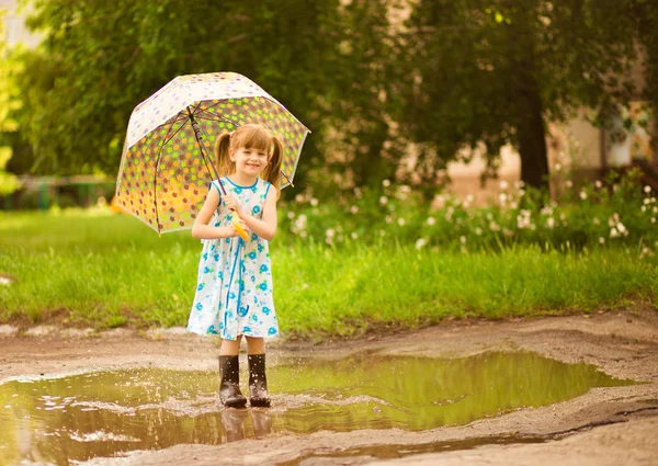 Gelukkig kind meisje in jurk met een paraplu en rubberen laarzen in plas op de zomer wandeling — Stockfoto