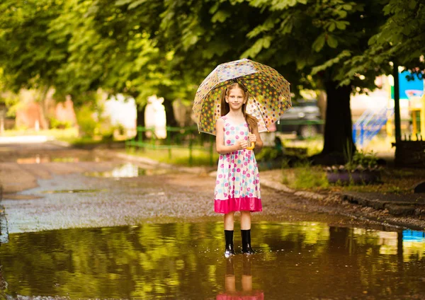Heureux enfant fille en robe avec un parapluie et bottes en caoutchouc dans la flaque d'eau sur la promenade d'été — Photo