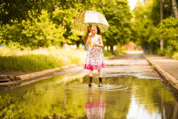 Happy funny kid girl with umbrella jumping on puddles in rubber boots and in polka dot dress and laughing — Stock Photo, Image