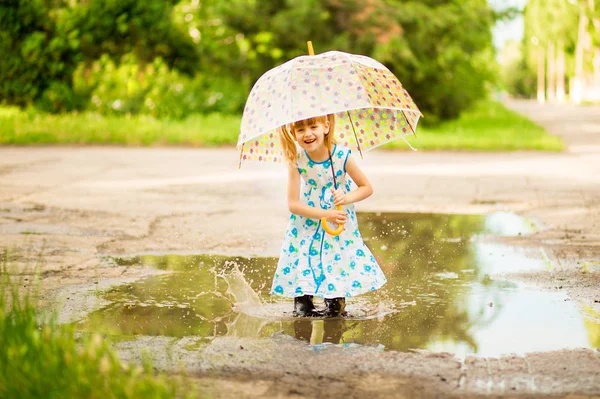 Menina garoto engraçado feliz com guarda-chuva pulando em poças em botas de borracha e em polka dot vestido e rindo — Fotografia de Stock