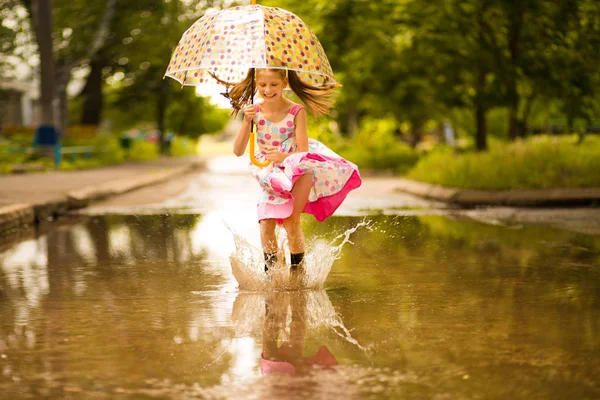 Menina garoto engraçado feliz com guarda-chuva pulando em poças em botas de borracha e em polka dot vestido e rindo — Fotografia de Stock