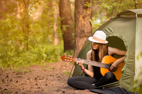 Chica turística en sombrero se sienta en la tienda y el concepto de tocar la guitarra. El turismo descansa en la naturaleza. Camping estilo de vida — Foto de Stock