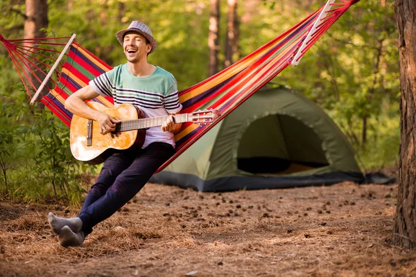 Real relaxation. Handsome young man in hat play guitar while lying in hammock , camping on background