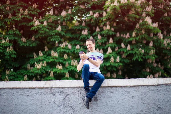 Homme souriant avec smartphone à la main assis dans la rue et en utilisant un messager. concept de réseau social — Photo