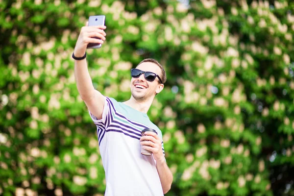 Cutie hombre en gafas de sol con taza de café mirando el teléfono móvil al aire libre — Foto de Stock