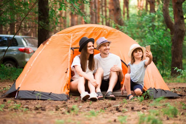 camping, hike, technology and people concept - happy family with smartphone taking selfie at campsite