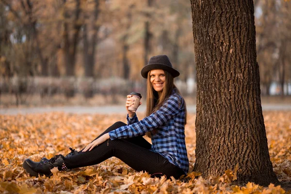 Koncepcja upadku. Szczęśliwa i wesoła Kobieta w kapeluszu, picie kawy siedząc na liście parku pod jesienią liści. Piękna młoda nowoczesna kobieta uśmiechnięta — Zdjęcie stockowe