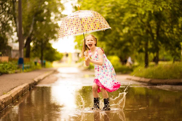 Feliz niña divertida con paraguas saltando sobre charcos en botas de goma y en vestido de lunares y riendo — Foto de Stock