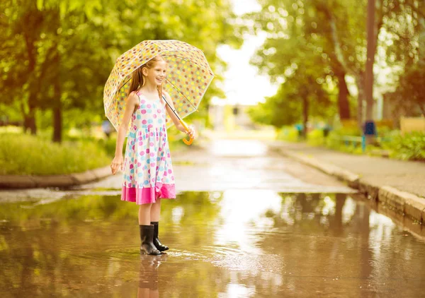 Felice ragazza divertente bambino con ombrello saltare su pozzanghere in stivali di gomma e in abito a pois e ridere — Foto Stock