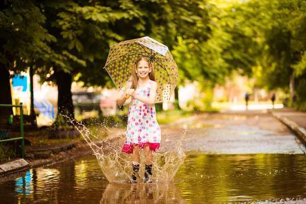 Happy funny kid girl with umbrella jumping on puddles in rubber boots and in polka dot dress and laughing — Stock Photo, Image