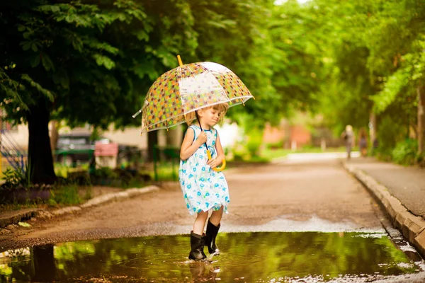 夏の散歩に水たまりで傘とゴムブーツを着たドレスを着た幸せな子供の女の子 — ストック写真
