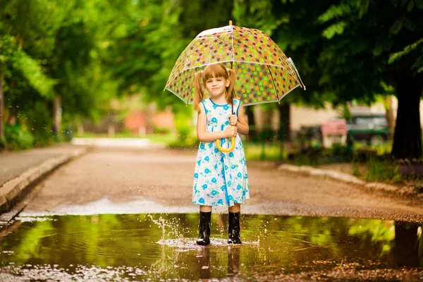 Happy child girl in dress with an umbrella and rubber boots in puddle on summer walk — Stock Photo, Image