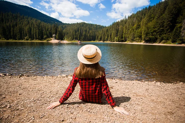 Turista mujer en sombrero sentado hacia atrás y viendo lago mágico en el paisaje del país montañoso . — Foto de Stock