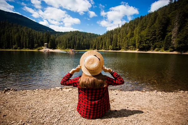 Back view of female tourist in hat with hands up enjoying freedom and amazing scenery of mountain lake. Young woman admiring lake and nature during trip