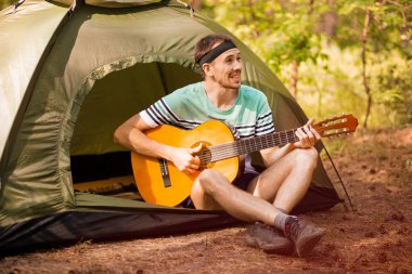 Mutlu genç adam kamp yapıyor ve ormanın günbatımına karşı dinlenmek için gitar enstrümantal bir müzik çalıyor. Yaban hayatı hayat tarzı konsepti.