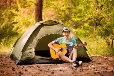 Mutlu genç adam kamp yapıyor ve ormanın günbatımına karşı dinlenmek için gitar enstrümantal bir müzik çalıyor. Yaban hayatı hayat tarzı konsepti.