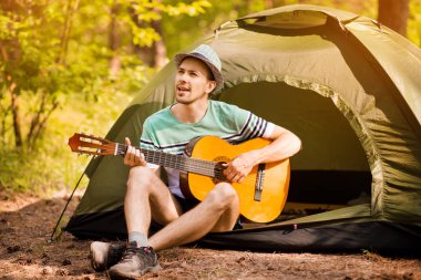 Mutlu genç adam kamp yapıyor ve ormanın günbatımına karşı dinlenmek için gitar enstrümantal bir müzik çalıyor. Yaban hayatı hayat tarzı konsepti.