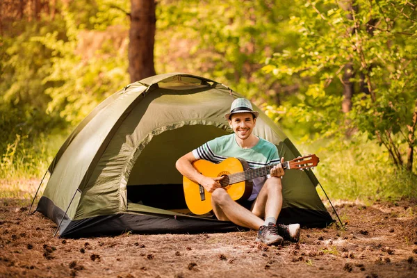 Feliz joven acampando y strum una guitarra instrumental de música para relajarse en el fondo de la puesta de sol del bosque. Wilderness Viajes concepto de estilo de vida . — Foto de Stock
