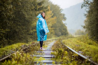 Kız Gezgin giyiyor mavi ceket ve go orman tren tarafından. Mavi yağmur ceket kız ormanda, sis yürür iken koyu yeşil tonları ile sonbahar, yağmur sezonu