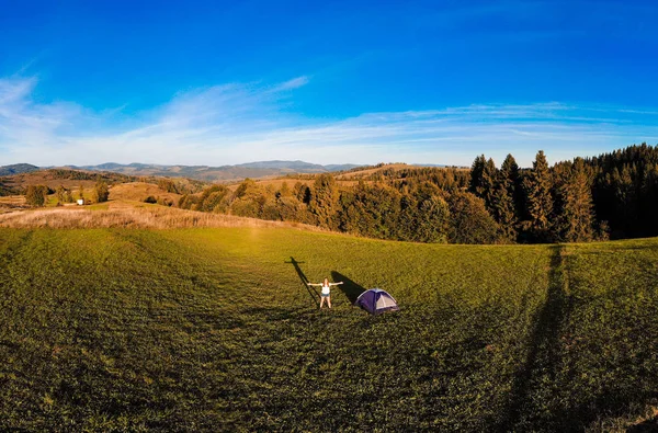Vista aérea panorámica de la mujer excursionista feliz con brazos abiertos permanecer cerca de la tienda de campaña alrededor de las montañas horizonte puesta de sol disfrutando del ocio y la libertad . —  Fotos de Stock
