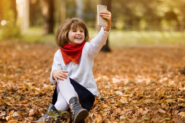 Niña divertida con una gran sonrisa tomando selfie usando teléfono inteligente al aire libre en el parque de otoño. Feliz infancia. — Foto de Stock