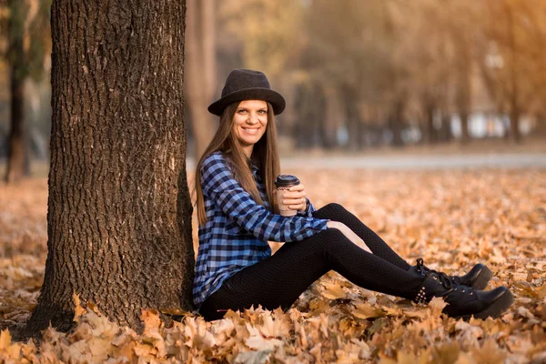 Happy woman in black hat drinking hot coffee in park under fall foliage. Beautiful young modern smiling woman on coffee break . Fall concept