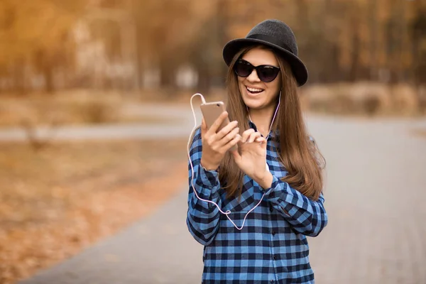 Mujer joven belleza en camisa azul sombrero negro y gafas de sol mensaje de escritura en el teléfono celular mientras camina por la calle de otoño — Foto de Stock