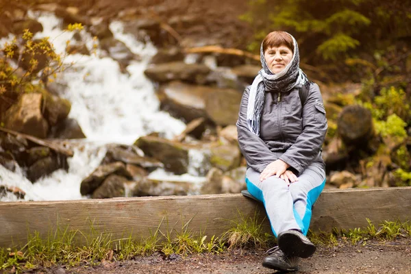 Vacker senior kvinna med halsduk runt huvudet från kall poserar nära skogen vattenfall — Stockfoto