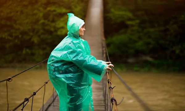 Viajero senior hermosa mujer en chaqueta de lluvia azul cruzar el río por un puente abisagrado en el bosque, disfrutando del silencio y la armonía de la naturaleza —  Fotos de Stock