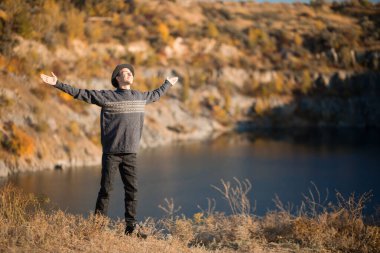 Özgürlük kavramı. Günbatımında kolları açık siyah şapkalı ve kazaklı özgür bir adam sonbahar gölü manzarasına hayran. Başarı. Seyahat et. İyi bir hayat..