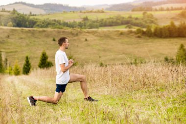 Koşucu, koşu parkurundan önce bacaklarını açıyor. Formda erkek koşucu, güzel sonbahar dağ manzarasında açık havada egzersiz yapıyor.