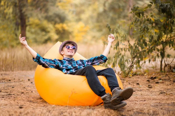 Pemuda tampan bertopi dan kacamata hitam berbaring di sofa karet dengan tangan ke atas dan menikmati waktu luang di udara terbuka sambil beristirahat di rumput di hutan musim gugur — Stok Foto