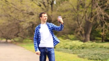 Genç adam selfie çekmek için telefon kullanıyor, parkta yürüyor ve fotoğraf ya da video için mobil uygulama kullanmaktan mutlu oluyor..